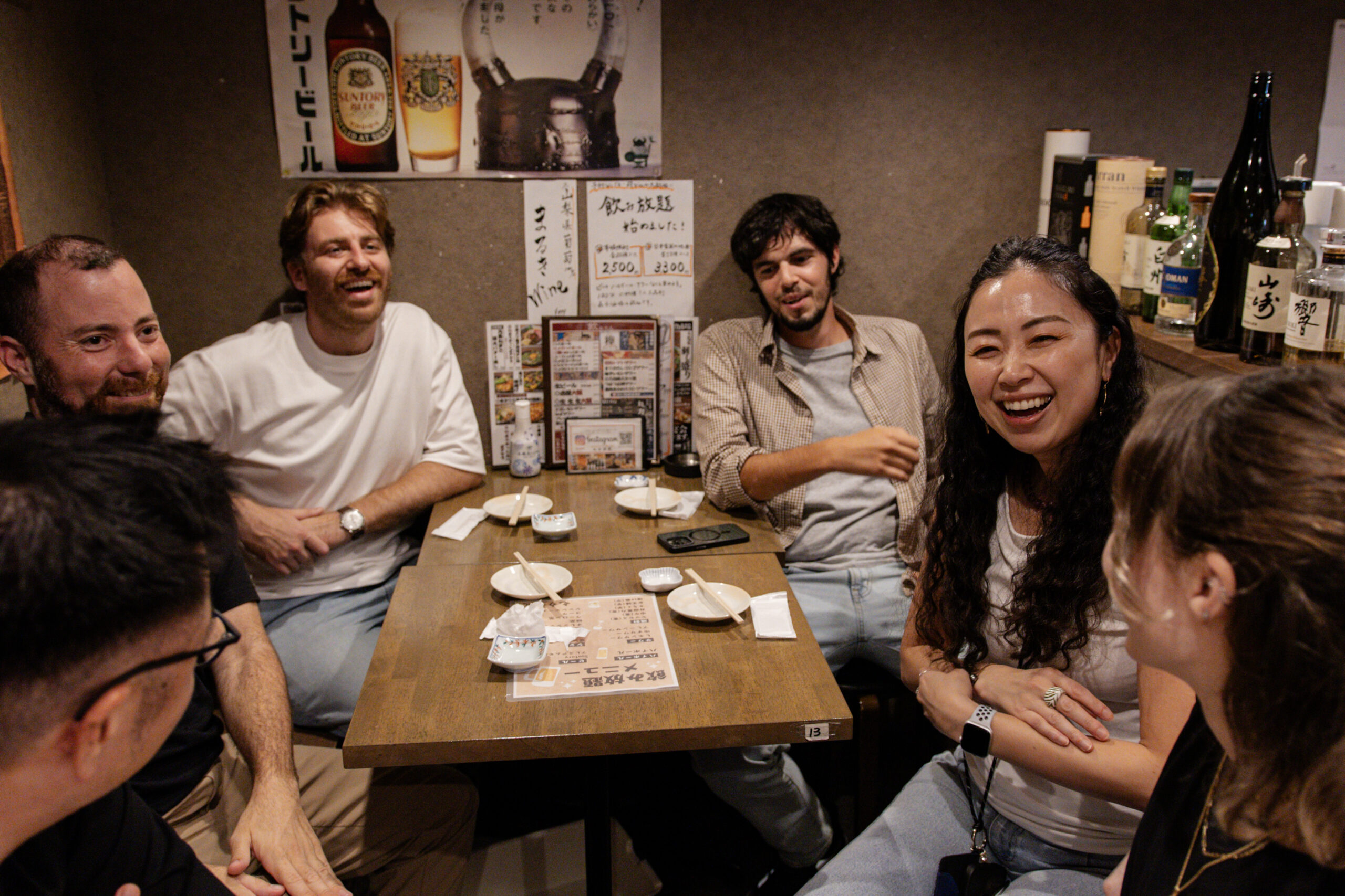 International participants chatting and laughing at an izakaya, with sake labels covering the wall behind them